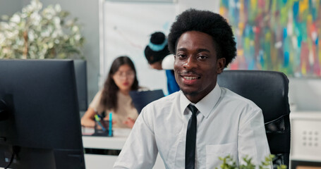 A joyful employee sits at his desk, focused on his computer screen, filling out forms, developing a new project, and looking at the camera with a big smile.
