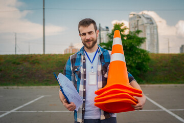 Smiling Caucasian man working in driving school, holding traffic cones and clipboard, standing...