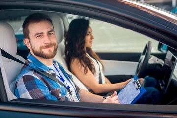 Young woman driving a car with male instructor. Lady looking on the road, man holding test drive...