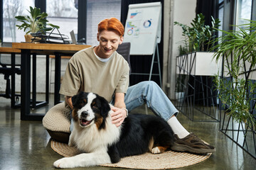 Young red haired man enjoys a joyful moment with his dog in a modern office space