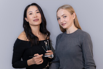 A professional makeup artist and a model pose together in a studio while holding beauty products. They are dressed stylishly, with a neutral background emphasizing elegance, professionalism