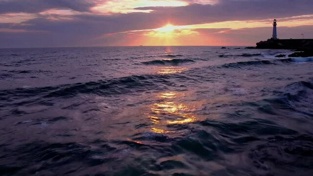 Dramatic sunset over ocean waves with lighthouse silhouette on rocky coastline