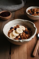 Fototapeta premium A bowl of crunchy granola with banana slices and blueberries sits on a wooden table, beside a rustic spoon and a side bowl of dried fruits, making for a warm, inviting breakfast Generative AI