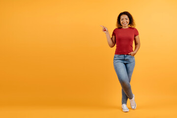 Full-length portrait of a smiling young African American woman pointing sideways, casually leaning on one leg, wearing a red shirt and jeans, vibrant yellow studio background with copy space