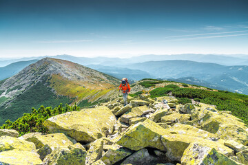 Naklejka premium Bearded backpacker in an orange jacket trekking over large boulders on a rugged mountain ridge. Panoramic mountain views in the background.