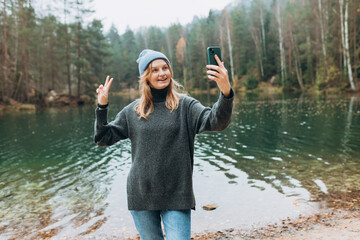 Happy blonde Woman makes selfie mobile photo enjoying mountain lake landscape. Nature freedom adventure, vacation video call, girl tourist outdoors. Concept of an active lifestyle