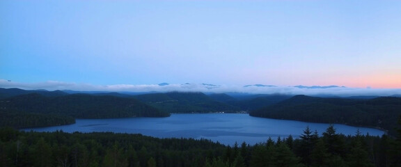 A panoramic view capturing a calm lake, bordered by dense forests and mist-covered hills, under the twilight sky. The peaceful ambiance evokes feelings of tranquility and appreciation of nature's beau