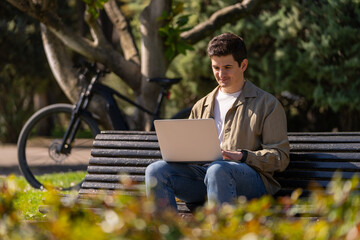 Fototapeta premium Chico escribiendo en portatil en un banco en el parque un dia soleado con una bici electrica