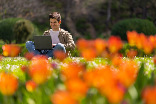 Joven caucasico sonriente en el parque sentando en el cesped trabajando con portatil negro un dia con sol