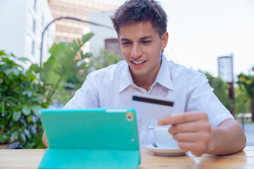 caucasian young businessman uses a credit card and tablet to make an online payment while sitting outdoors. He smiles during the digital transaction at a table in a relaxed urban setting.