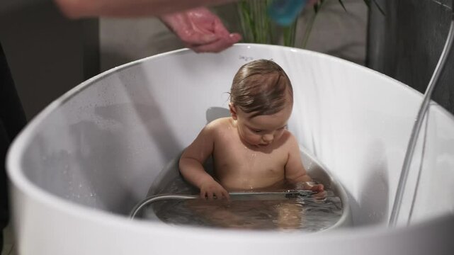A loving father gently shampoos his 1 year old son&rsquo;s head in the bathtub while the boy joyfully plays with the shower, creating a heartwarming and playful bath time scene