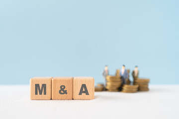 M and A, Mergers and Acquisitions, text on wooden cube blocks with blurred stack of coins and...