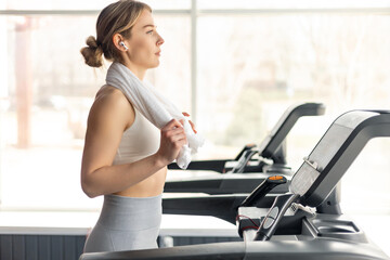 Fototapeta premium Happy European woman in sportswear and wireless headphones and towel around neck doing hard cardio workout on treadmill, lady working out in empty gym
