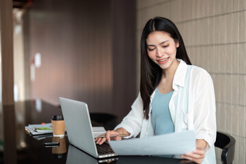 Remote Work and Focused Productivity. A woman smiles as she reviews documents while working on her laptop at a stylish workspace.
