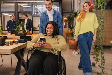 Smiling businesswoman using smartphone in wheelchair at modern office with colleagues