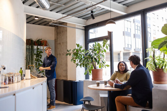 Colleagues chatting in modern office break room, man washing dishes