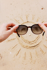 Hands holding sunglasses with smiley face drawn in sand in front of person wearing sunglasses on...