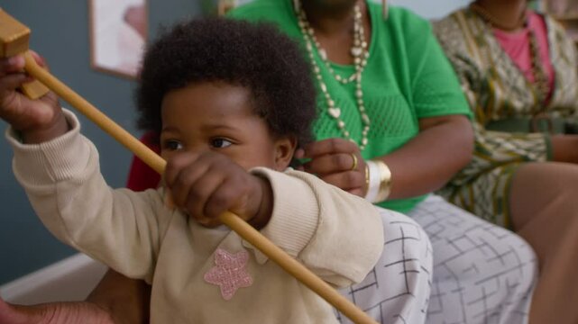 African American toddler playing with wooden toy, while her mother and aunt looking after her, sitting on couch in living room