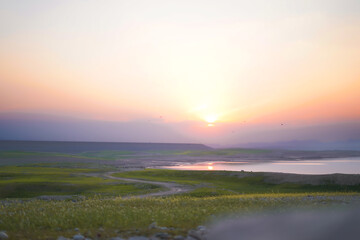 Obraz premium A breathtaking sunset over lush green fields, a winding dirt path, and a calm reflective lake, creating a peaceful and scenic view.