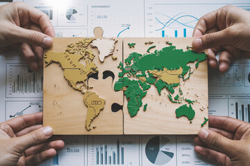 Wooden world map puzzle pieces, beige and green, held by hands above business graphs, showcasing global connection and collaboration