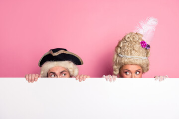 Two individuals in vintage aristocratic costumes peeking over a white board with a pink background