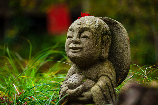 A joyful Warabe-Jizo statue cradling a child, decorated with a red maple leaf, amidst vibrant autumn grass and leaves, Kyoto, Honshu