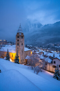 Church of Saint Pantalon, snow covered town centre and mountainous background in winter at dusk, Courmayeur, Aosta Valley, Italian Alps