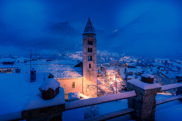 Church of Saint Pantalon and snow covered town centre and mountainous background in winter at dusk, Courmayeur, Aosta Valley, Italian Alps