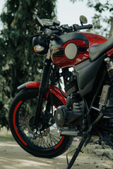 A red and black classic motorcycle with a retro design, round gauges, and spoked wheels, parked on a dirt path with trees in the background.