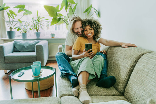 Multiracial young couple watching smart mobile phone sitting on the sofa at home - Happy diverse husband and wife downloading mobile app  - Technology life style concept