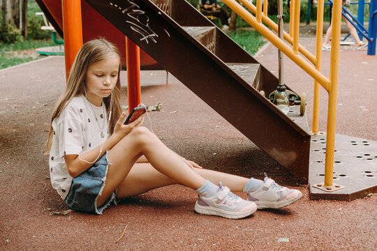 A teenage girl is looking at her smartphone. She is sitting on the ground in the sports field checking her phone. Internet addiction among kids and loneliness among teenagers - Powered by Adobe