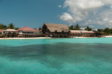 Crystal clear turquoise water gently laps against the shore in front of a traditional thatched roof restaurant in zanzibar, tanzania, creating a serene tropical paradise