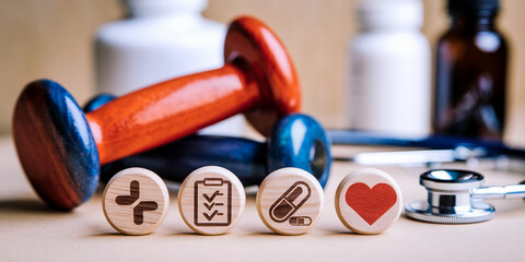 Wooden discs with health icons; cross, checklist, pill, heart; near dumbbells and stethoscope, symbolizing wellness, healthcare, and healthy lifestyle choices