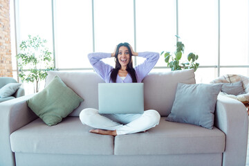 Fototapeta premium Happy woman sitting on a cozy sofa with a laptop in a modern bright living room during the day enjoying a relaxed moment