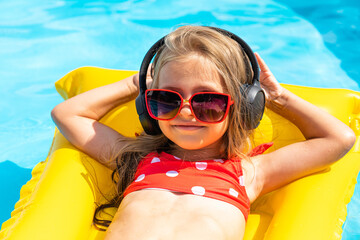 Happy little girl lying on inflatable mattress in swimming pool and listening to music via...