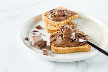 Spreading sweet chocolate cream, cashew, chocolate chunks onto toast on table.