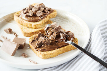 Spreading sweet chocolate cream, cashew, chocolate chunks onto toast on table.