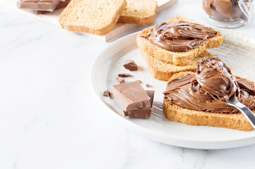 Spreading sweet chocolate cream, cashew, chocolate chunks onto toast on table.