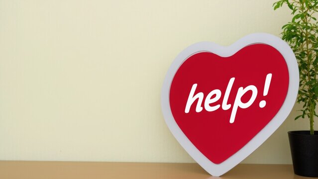 A heart-shaped sign reading "help!" in white text on a vivid red background lies on a wooden surface next to a small potted plant, symbolizing support, compassion, and urgent assistance