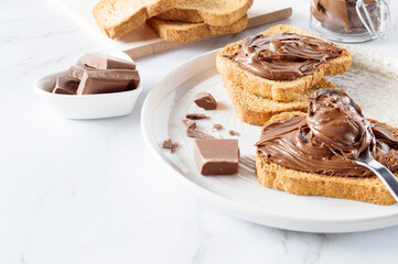 Spreading sweet chocolate cream, cashew, chocolate chunks onto toast on table.