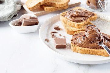 Spreading sweet chocolate cream, cashew, chocolate chunks onto toast on table.