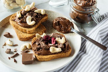 Spreading sweet chocolate cream, cashew, chocolate chunks onto toast on table.