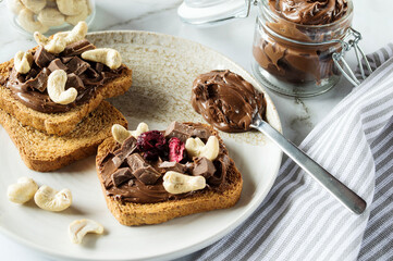 Spreading sweet chocolate cream, cashew, chocolate chunks onto toast on table.