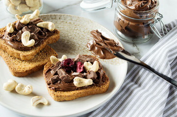 Spreading sweet chocolate cream, cashew, chocolate chunks onto toast on table.