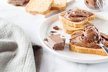 Spreading sweet chocolate cream, cashew, chocolate chunks onto toast on table.