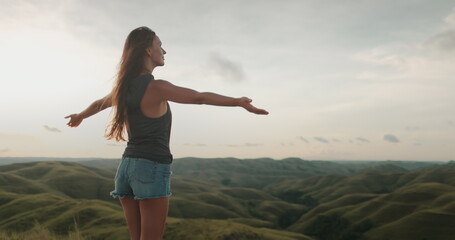 Young woman is embracing the beauty of nature, raising her arms towards the sky on top of a hill with a valley on the background