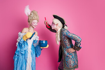 Playful couple in vibrant baroque costumes against a pink backdrop, captured mid-action during a masquerade theme party