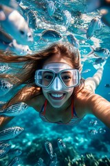 Fototapeta premium A gorgeous and sexy happy mixed-race young woman with curly hair in a swimming mask and fins snorkeling among the corals. Exotic beach getaway. A dense school of silver fish swims next to her.