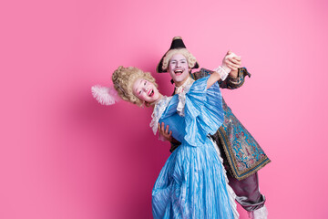 Joyful couple dressed in elegant historical costumes poses against a pink backdrop, embodying love and nobility