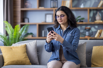 Young Indian woman in shirt and glasses sitting on sofa at home and using mobile phone